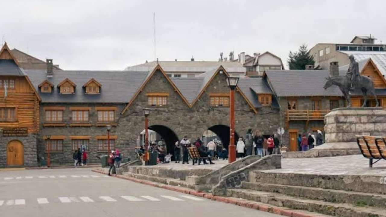 Praça do Centro Cívico de Bariloche com construções de pedra e madeira no estilo alpino, estátua e visitantes caminhando.