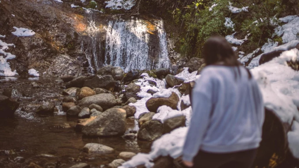 Cachoeira em um riacho de montanha cercada por pedras e neve derretendo, com uma pessoa caminhando ao fundo.