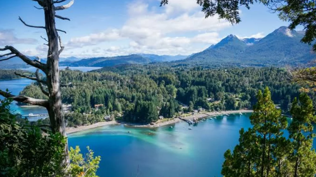 Vista panorâmica de Villa La Angostura, mostrando o Lago Nahuel Huapi cercado por montanhas verdes e o céu azul ao fundo. Um cenário típico da Patagônia com águas cristalinas e vegetação abundante.