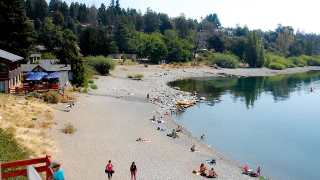 Playa Bonita em Bariloche, com faixa de areia/cascalho, pessoas tomando sol e vegetação nas margens.