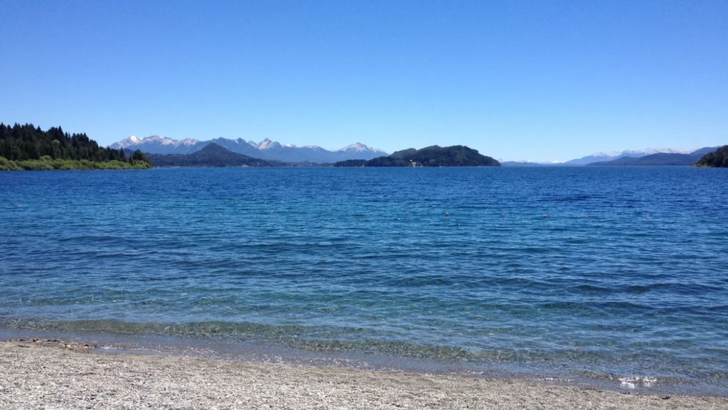 Vista panorâmica de um lago cristalino rodeado por montanhas, sob um céu azul claro, em Bariloche.