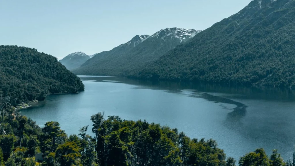 Lago rodeado de montanhas cobertas de florestas densas, com águas tranquilas refletindo o céu azul. A paisagem remete à tranquilidade e à beleza natural da região da Patagônia.