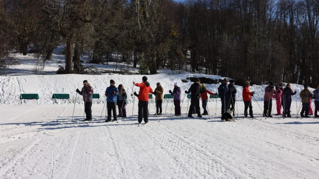 Grupo de pessoas participando de uma aula de esqui em uma pista plana, com árvores ao fundo e um cachorro sentado na neve observando.