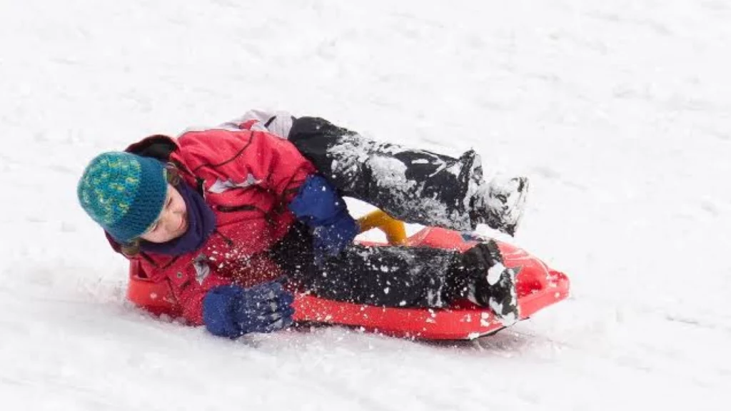 Criança deslizando na neve em um trenó vermelho, vestindo um casaco e gorro, coberta de neve enquanto se diverte.