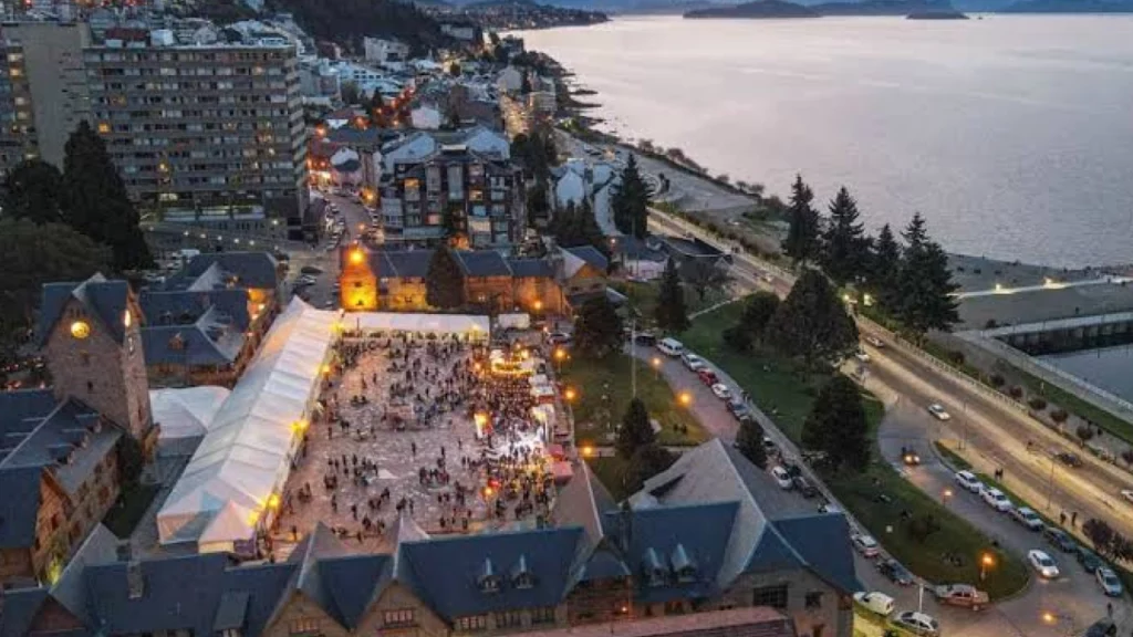 Vista aérea do centro cívico de Bariloche ao pôr do sol, com o lago Nahuel Huapi ao fundo e um grande festival acontecendo na praça central.