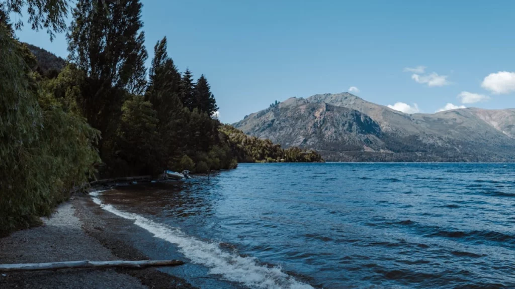 Orla tranquila do lago Nahuel Huapi, cercada por árvores e montanhas ao fundo.