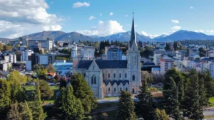 Vista aérea da Catedral de Bariloche, cercada por árvores, com a cidade e as montanhas cobertas de neve ao fundo, em um dia ensolarado.