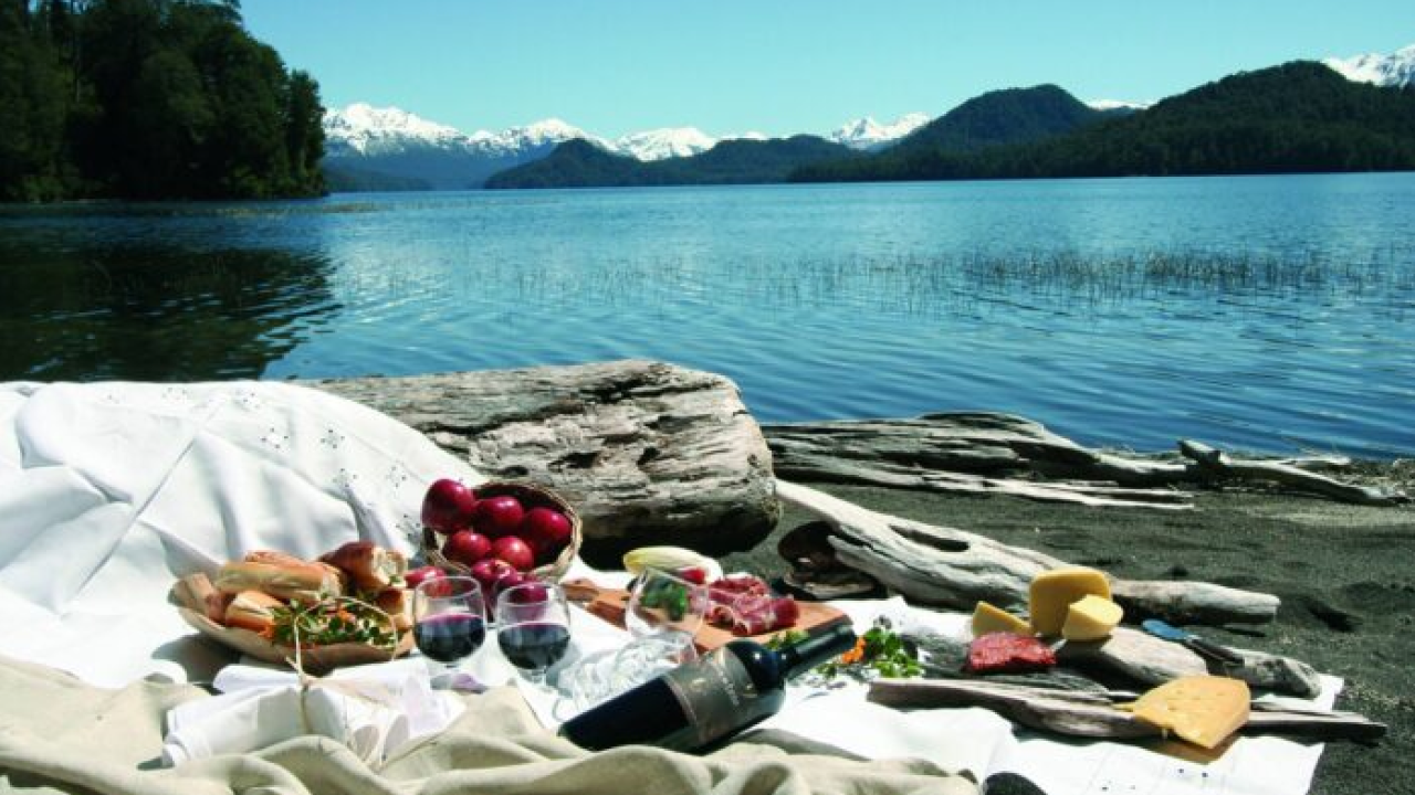 Piquenique à beira de um lago em Bariloche, com tábuas de frios e taças de vinho, montanhas nevadas ao fundo.