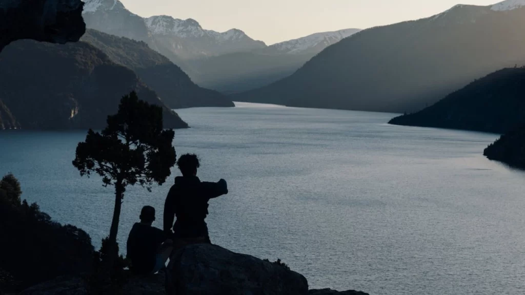 Silhuetas de duas pessoas admirando o lago Nahuel Huapi ao entardecer, com montanhas e neve ao fundo.