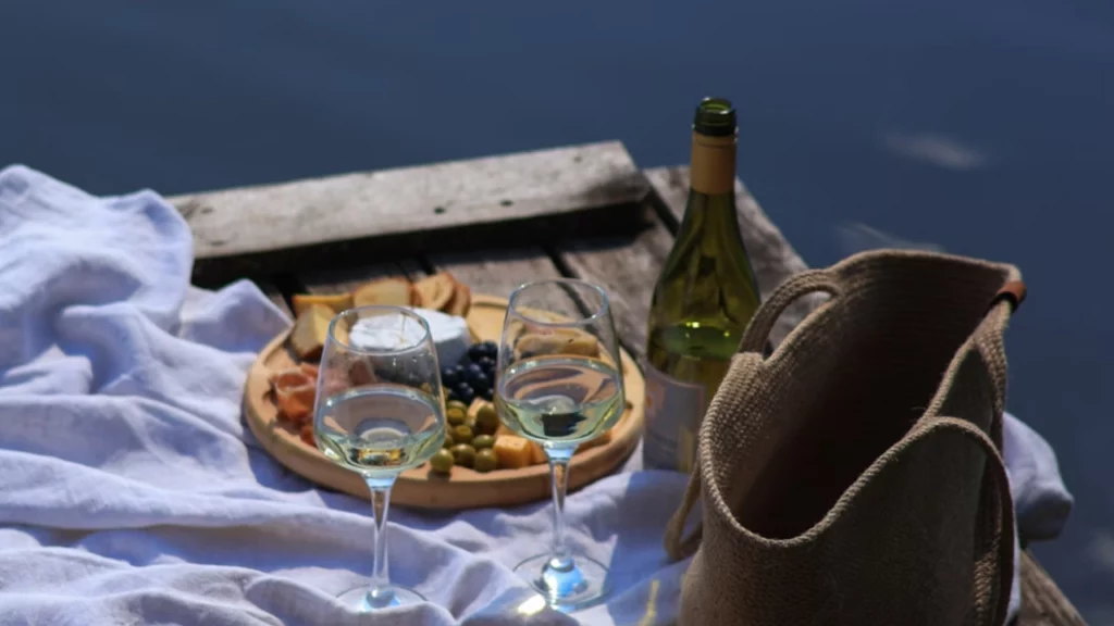 Uma cena de piquenique ao lado de um lago, com duas taças de vinho branco, uma garrafa de vinho, um chapéu de palha e uma cesta sobre uma toalha branca. Ao fundo, vê-se a superfície calma da água, refletindo o céu azul.