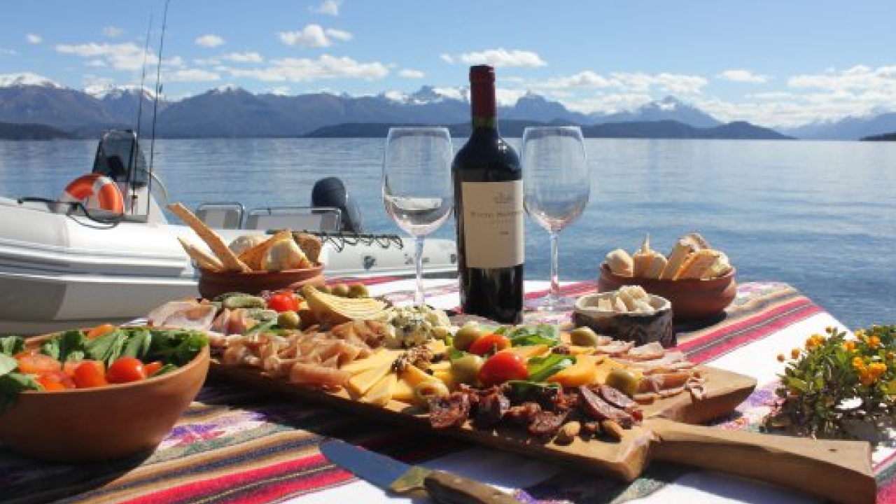 Mesa de piquenique com vinho e tábuas de frios à beira do lago em Bariloche, barco ancorado ao fundo.