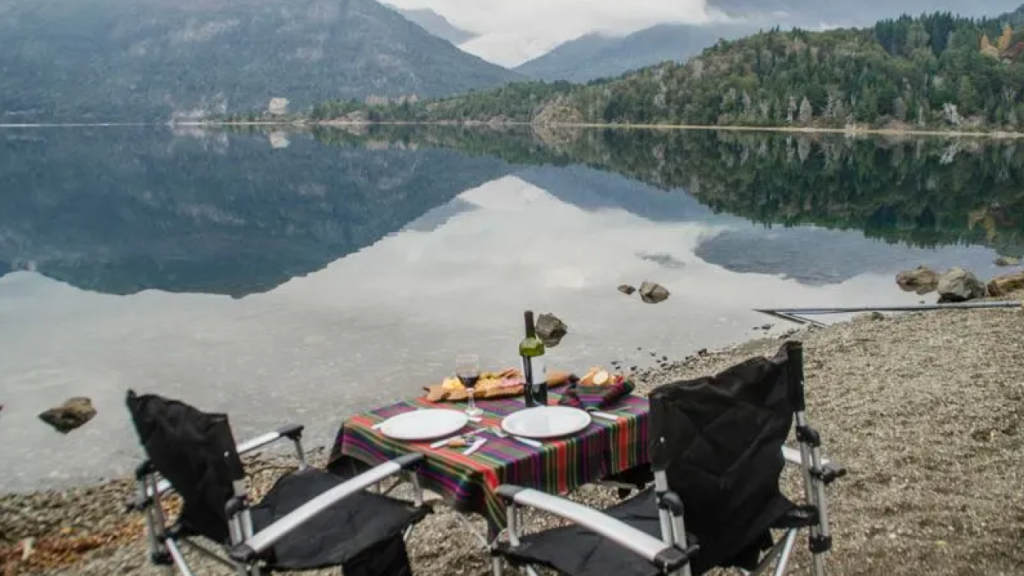 Duas cadeiras e mesa com lanche à beira de lago espelhado na Patagônia, montanhas refletidas na água.