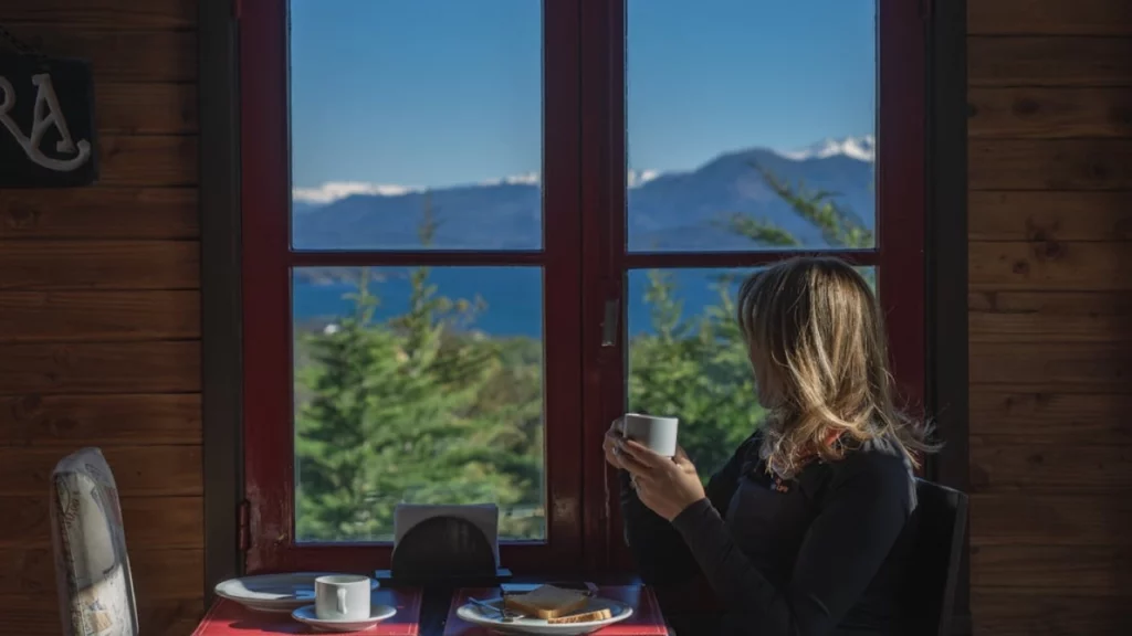 Vista de uma mulher contemplando o cenário de montanhas e lago em Bariloche, enquanto toma café junto à janela de uma cabana aconchegante.