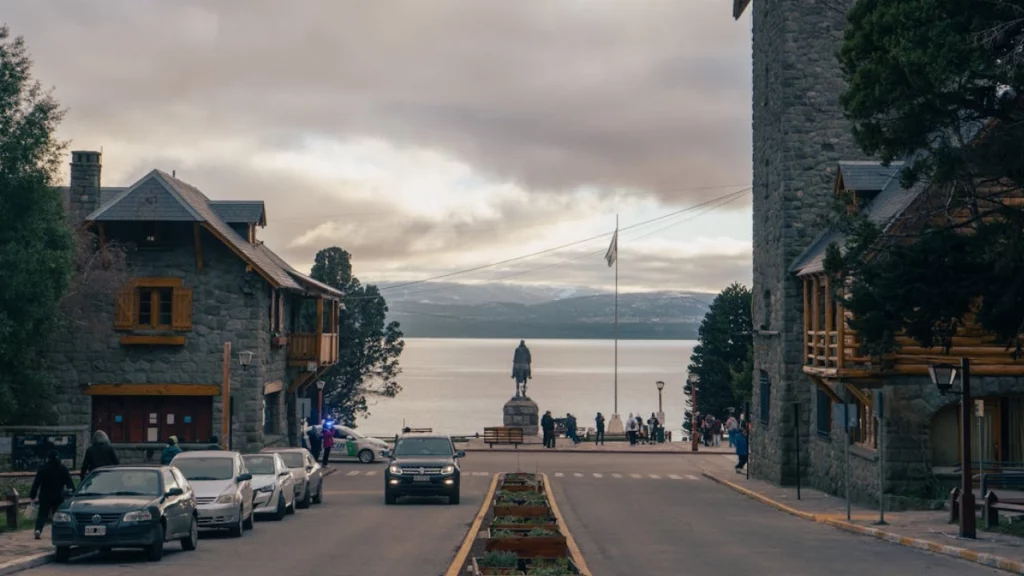 Rua central de Bariloche, com arquitetura rústica de pedra e madeira, e uma estátua com vista para o lago ao fundo.