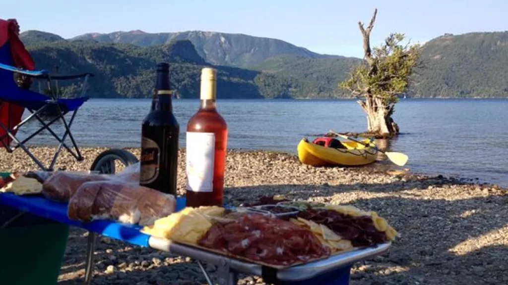 Tábua de queijos e embutidos com garrafas de vinho na praia de cascalho de um lago em Bariloche, caiaque ao fundo.