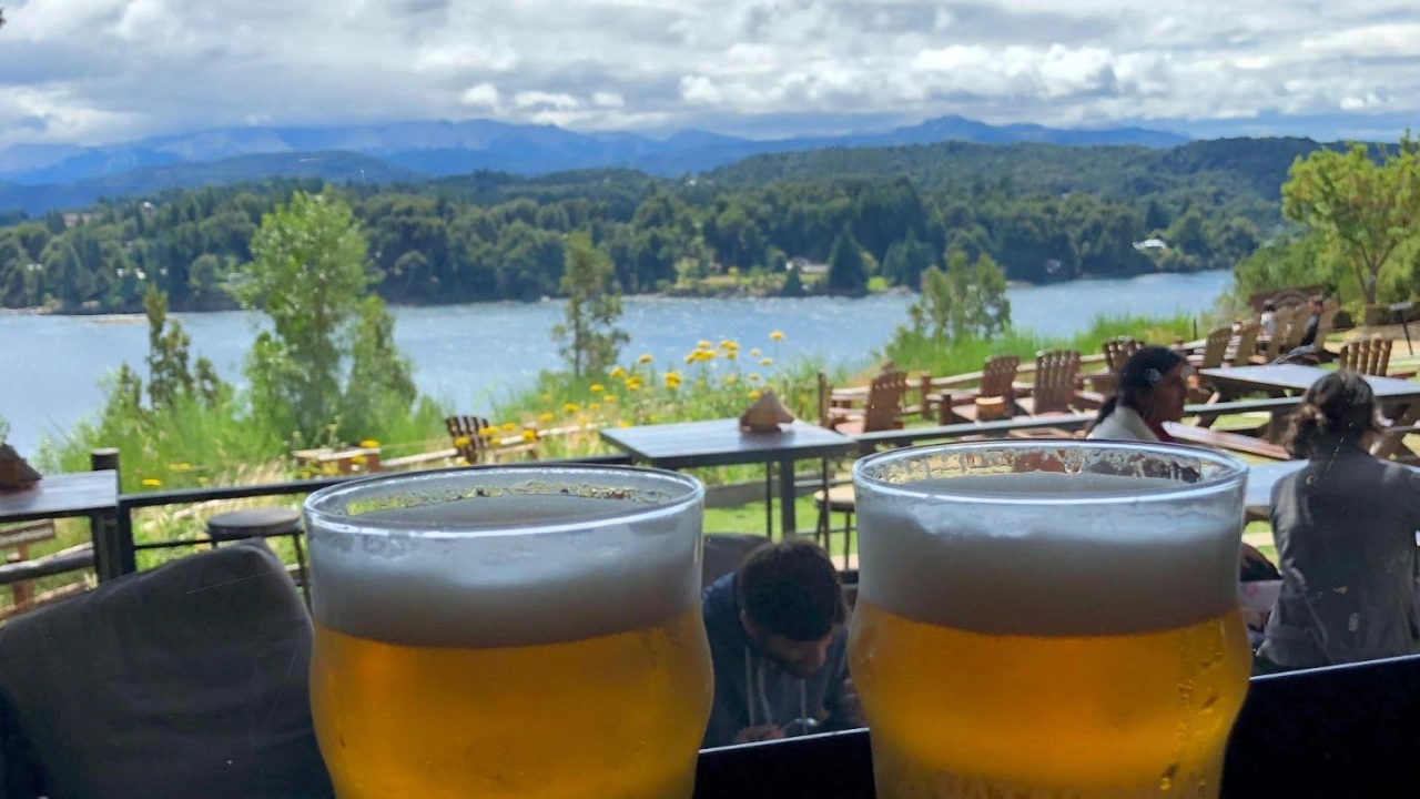 Dois copos de cerveja artesanal em uma cervejaria com vista para um lago e montanhas ao fundo em Bariloche, Argentina, em um dia ensolarado.