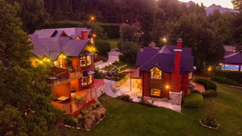 Complexo de chalés em Bariloche ao anoitecer, com luzes amarelas aconchegantes iluminando as varandas e jardins, rodeados por vegetação e montanhas ao fundo.