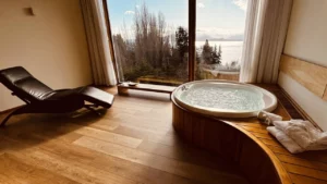 Jacuzzi em um quarto com uma grande janela que oferece uma vista panorâmica do Lago Nahuel Huapi, ao lado de uma cadeira de descanso em madeira e couro.