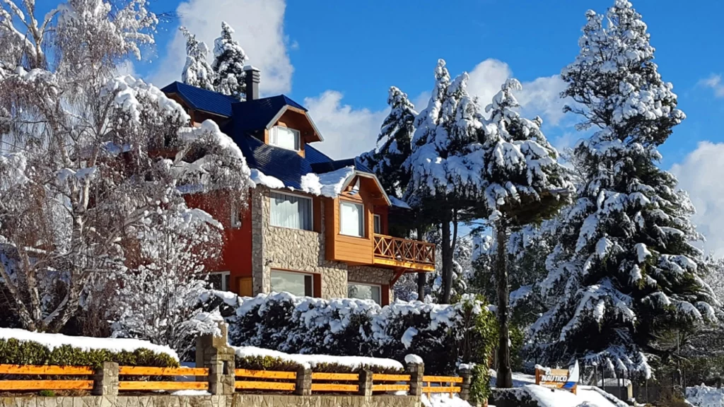 Chalé em Bariloche coberto de neve, cercado por árvores nevadas, destacando uma arquitetura charmosa de madeira e pedra com um céu azul claro ao fundo.