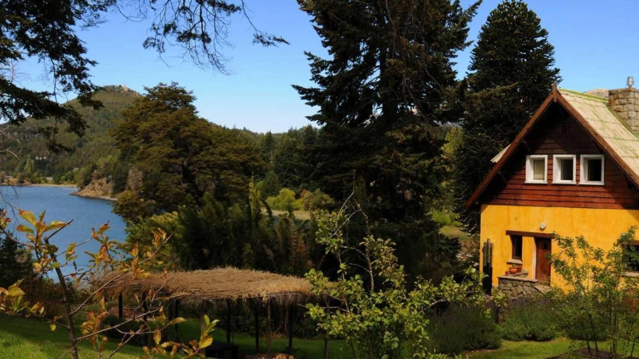 Casa de campo rústica em Bariloche, próxima ao Lago Nahuel Huapi, rodeada por vegetação e com uma vista parcial do lago, em meio a uma paisagem natural exuberante.