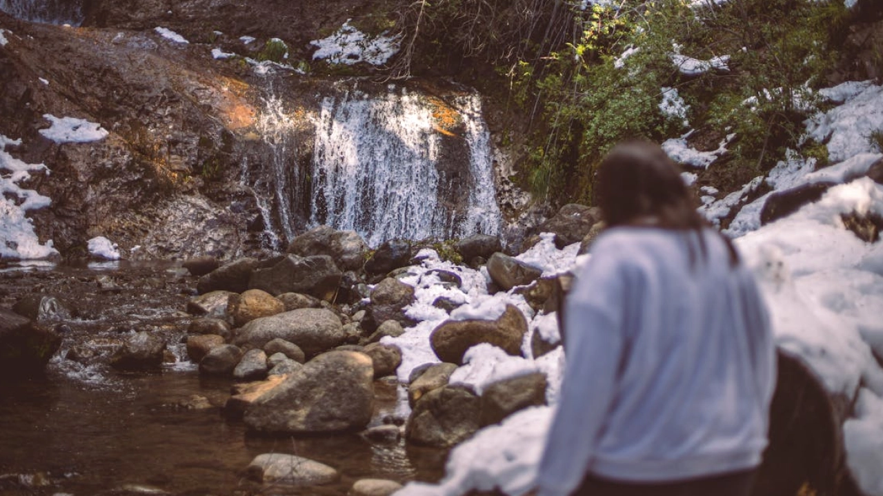 Trilha ao redor do Lago Gutiérrez, em Bariloche, com uma pequena cascata cercada por neve e pedras, ideal para momentos tranquilos em contato com a natureza.