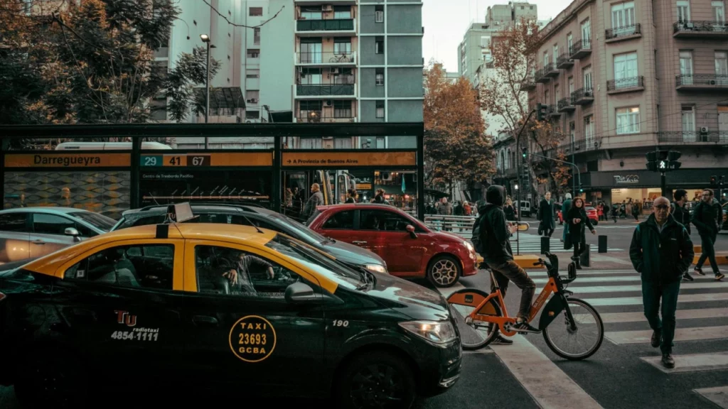 Cena urbana de Buenos Aires com táxis amarelos e pretos, ciclista em uma bicicleta laranja e pedestres cruzando a rua.