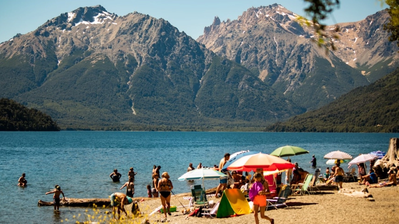 Praia movimentada em Bariloche, com turistas relaxando sob guarda-sóis coloridos e nadando no lago, enquanto montanhas nevadas formam o cenário ao fundo.