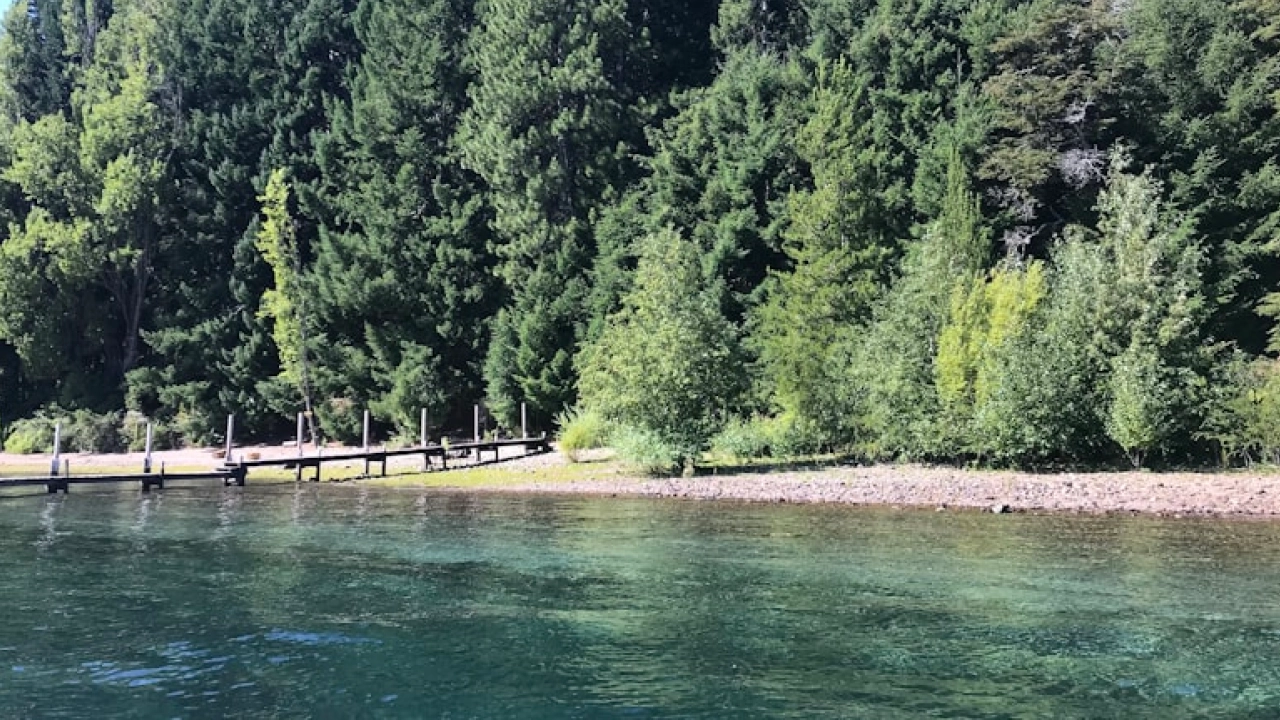 Pequena praia escondida em Bariloche, rodeada por uma floresta densa e com um píer de madeira à beira das águas cristalinas do Lago Moreno.