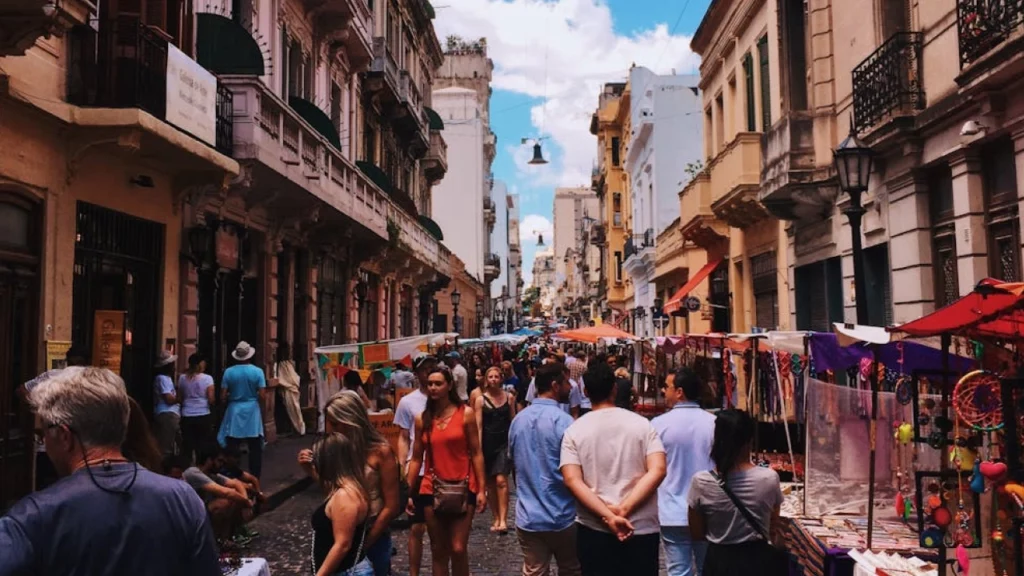 Rua movimentada em Buenos Aires com feirantes e turistas explorando o mercado de San Telmo, cercados por prédios históricos e cores vibrantes.