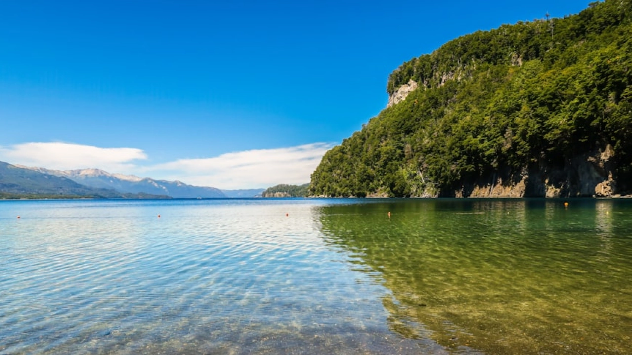 Praia de águas cristalinas em Bariloche, cercada por montanhas verdes e um céu azul intenso, refletindo a paisagem no Lago Nahuel Huapi.