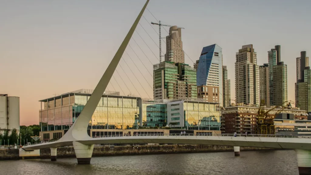 Uma vista do icônico Puente de la Mujer em Buenos Aires, Argentina, com modernos arranha-céus refletindo o pôr do sol no bairro de Puerto Madero. O rio e a estrutura arquitetônica elegante do ponte criam uma cena urbana sofisticada.