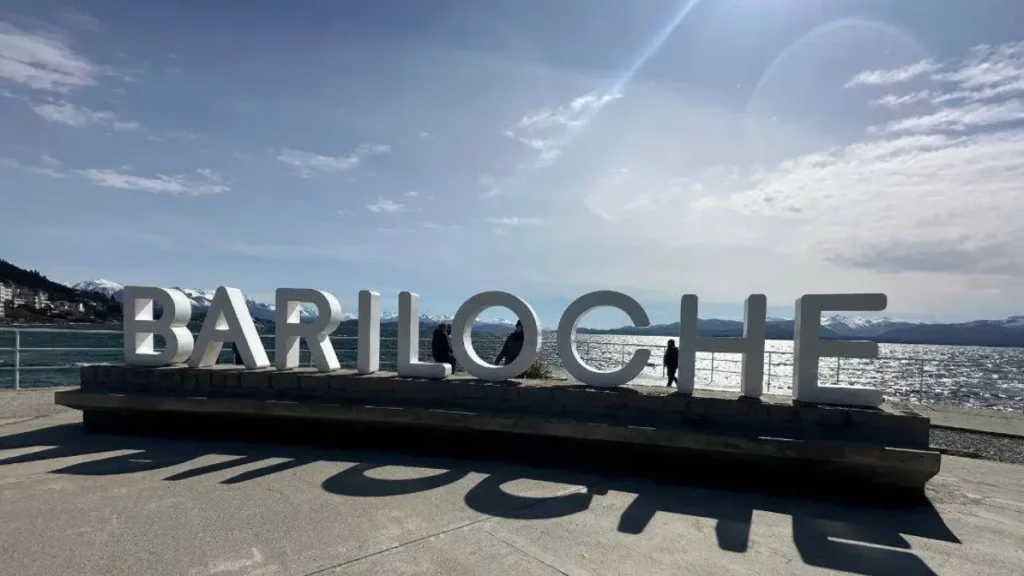 Una gran estructura con las letras en mayúsculas que conforman la palabra "BARILOCHE", ubicada junto a un lago y rodeada de montañas, con un cielo parcialmente nublado.