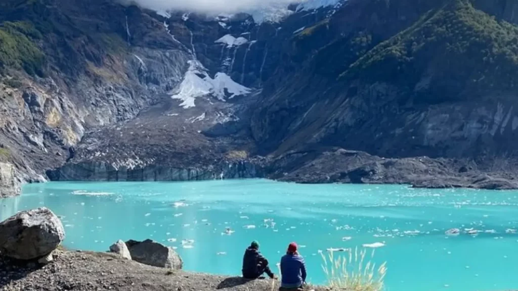 Duas pessoas sentadas em um terreno rochoso em frente a um lago de tom turquesa intenso, rodeado por encostas íngremes e geleiras, com parte do gelo derretido flutuando na água.