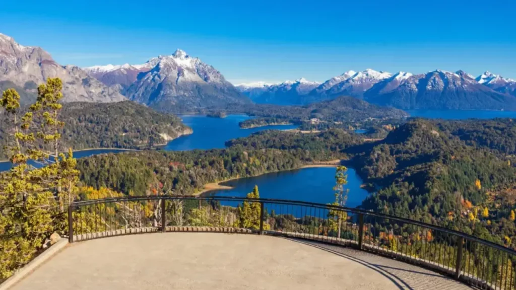 Vista panorâmica a partir de um mirante com guarda-corpo, mostrando vários lagos azuis entre florestas densas e montanhas nevadas ao fundo, em um dia de céu azul.