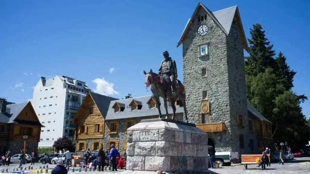 Estátua equestre em frente à torre de pedra do Centro Cívico de Bariloche em um dia ensolarado, com visitantes ao redor.