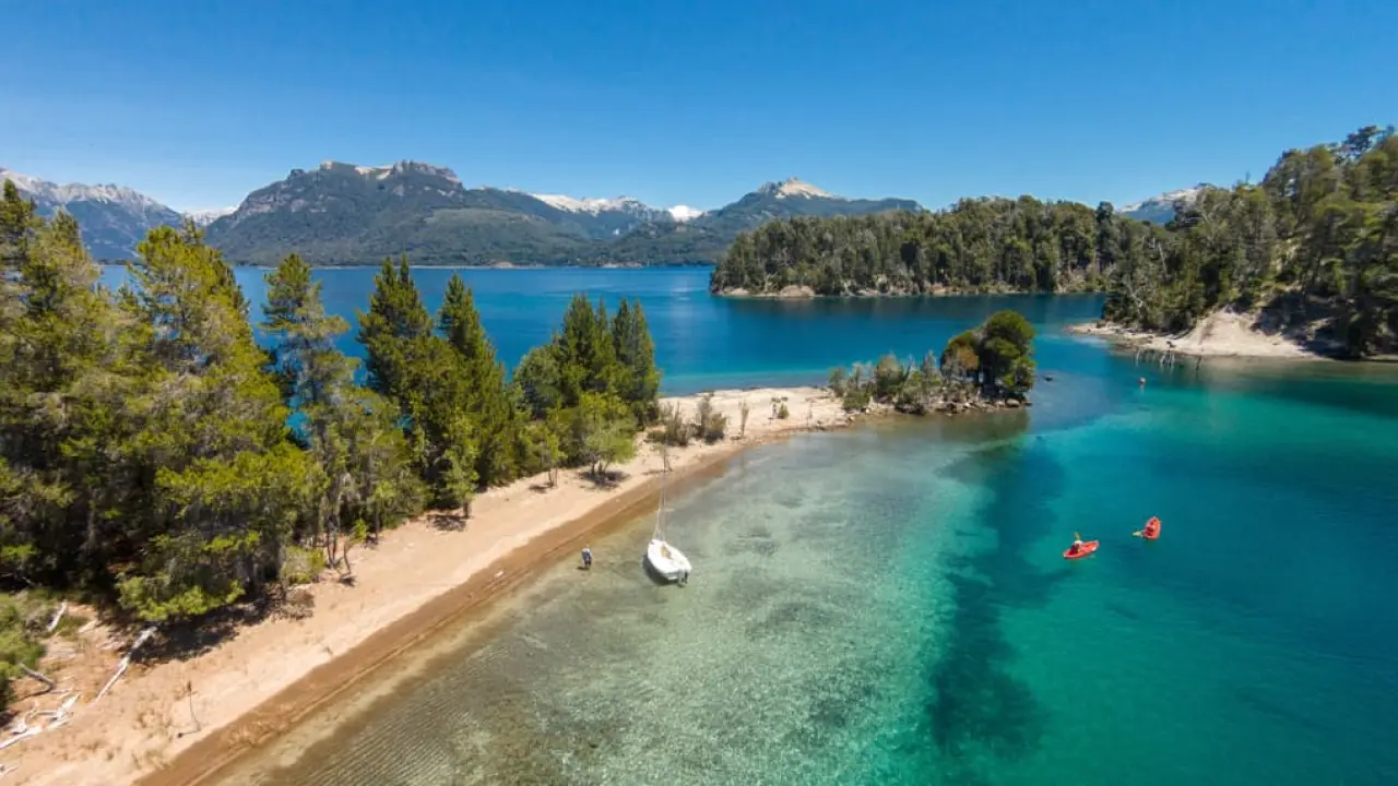 Uma pequena praia de areia clara em frente a águas cristalinas, onde há um barco ancorado e dois caiaques vermelhos. Florestas de pinheiros circundam o lago, com montanhas ao fundo sob céu azul.