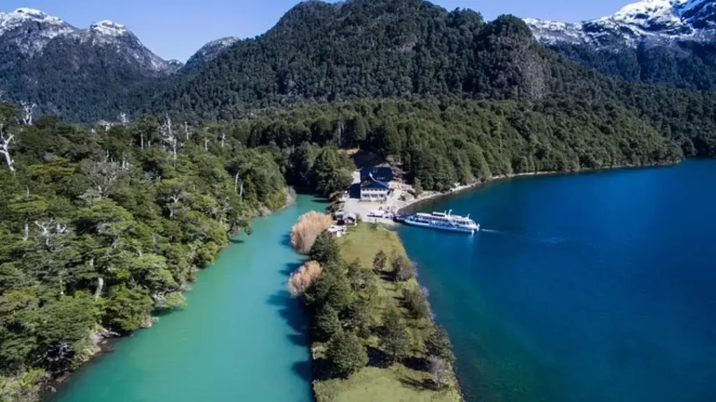 ista aérea de uma estreita península coberta por vegetação, com um edifício próximo à margem e um barco atracado em águas de cor turquesa. Ao fundo, montanhas cobertas por florestas e picos com neve.