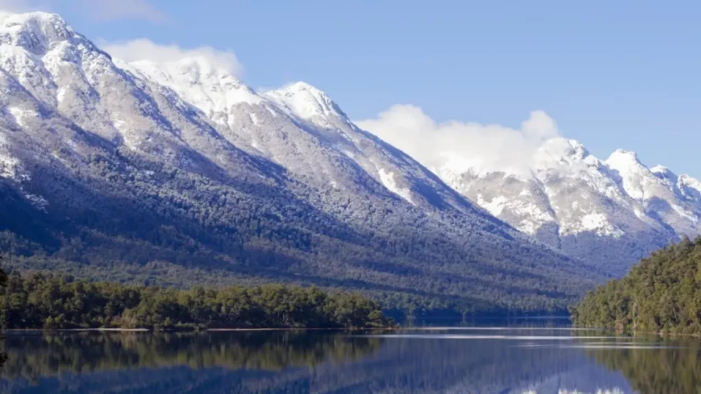 Montanhas altas cobertas de neve e floresta em suas encostas, refletidas em um lago calmo de cor azul profundo sob um céu claro.