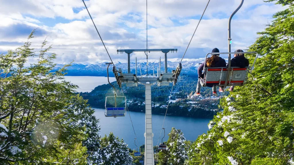 Teleférico subindo a montanha em Bariloche, com cadeiras transportando visitantes e o Lago Nahuel Huapi ao fundo em meio às montanhas.