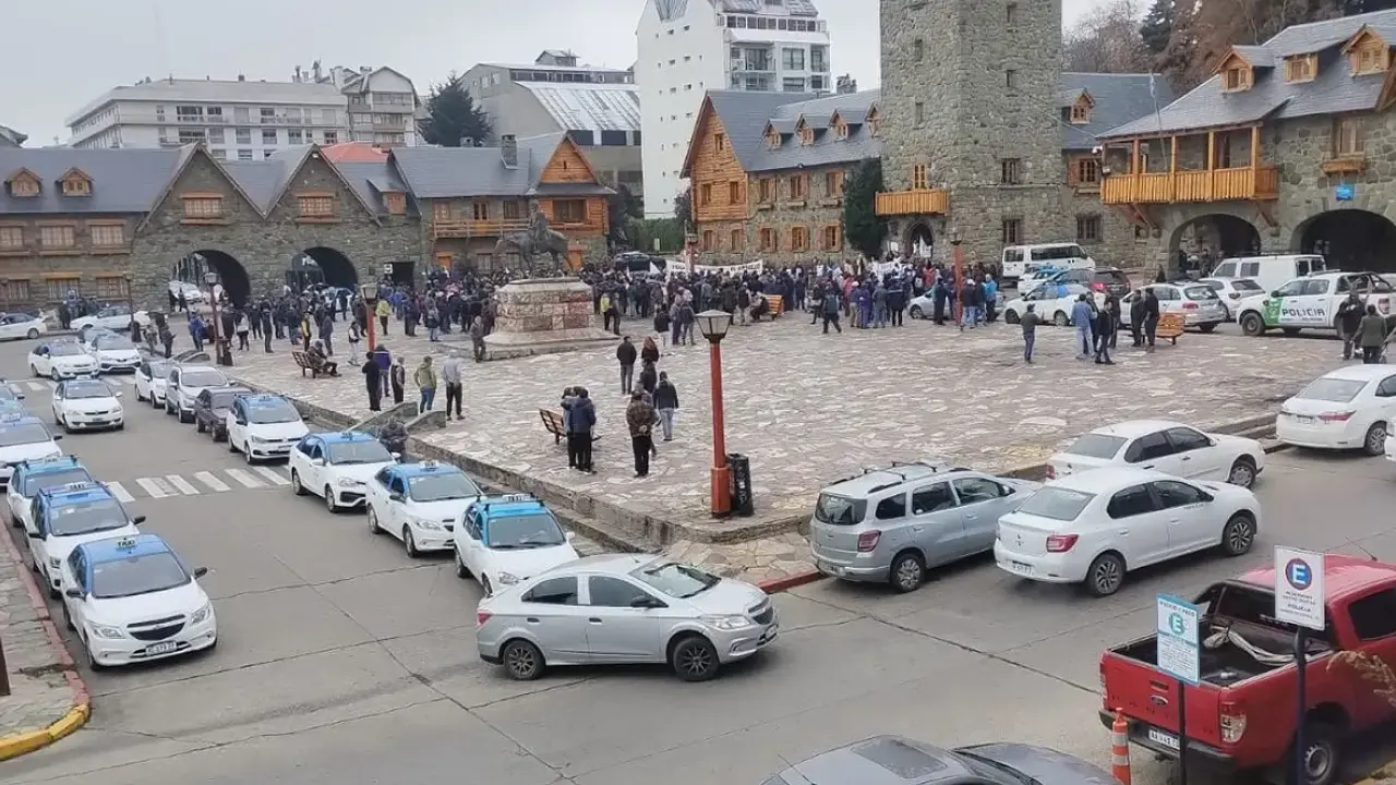 Centro Cívico de Bariloche com movimento de turistas, prédios de pedra e madeira em estilo alpino e fila de táxis estacionados.
