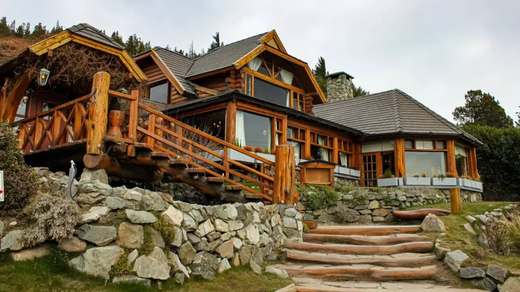Cabana em estilo rústico feita de madeira e pedra, com escada de troncos e janelas amplas, típica arquitetura de Bariloche.