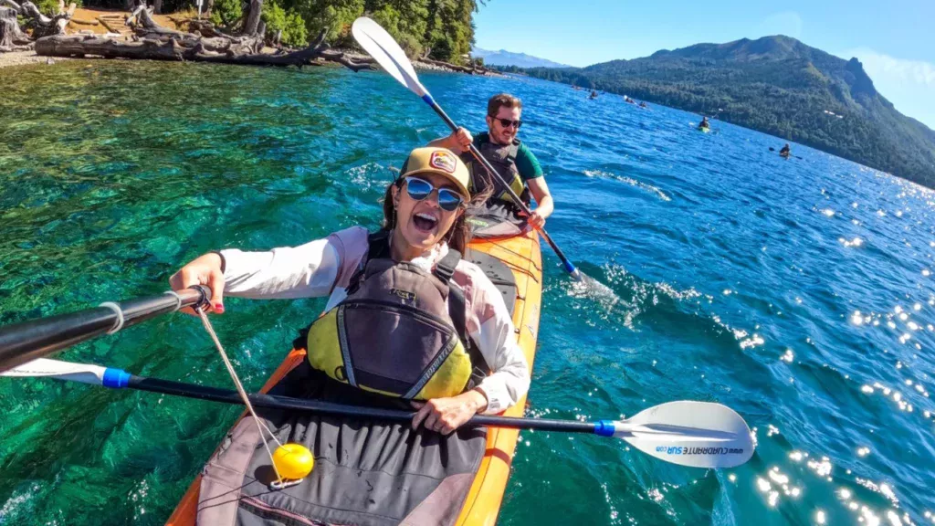 Casal remando em caiaque sobre águas cristalinas do Lago Gutiérrez, com montanhas ao fundo e céu aberto em Bariloche.