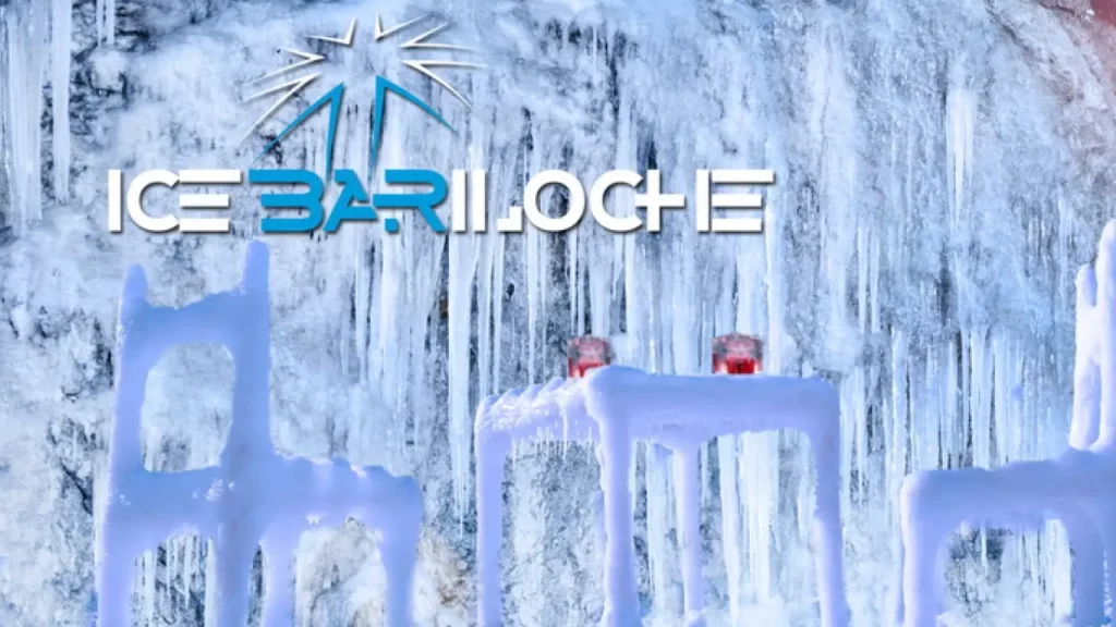   Logo do Ice Bar Bariloche sobre uma parede de gelo com estalactites congeladas. Em frente, cadeiras e uma mesa esculpidas em gelo com dois copos vermelhos sobre a mesa.
