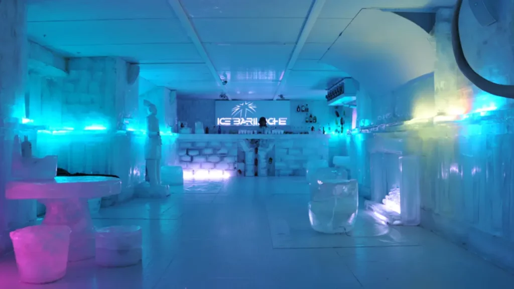 Interior iluminado do Ice Bar Bariloche, com mobiliário, esculturas e paredes feitas de gelo. Iluminação azul e roxa destaca o ambiente congelado, com o balcão de bar ao fundo.