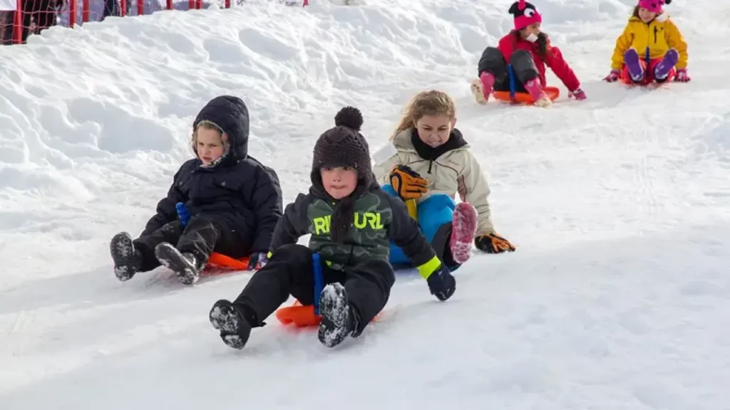 Crianças descendo uma pista de neve com pranchas de plástico, vestidas com roupas de inverno coloridas, em um ambiente recreativo cercado por grades vermelhas.