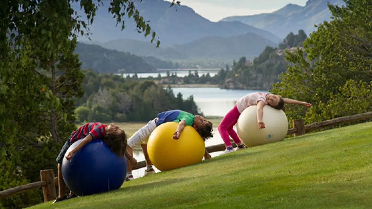 Três crianças deitadas sobre bolas de ginástica coloridas em um gramado inclinado com vista para um lago e montanhas ao fundo, em um dia ensolarado.
