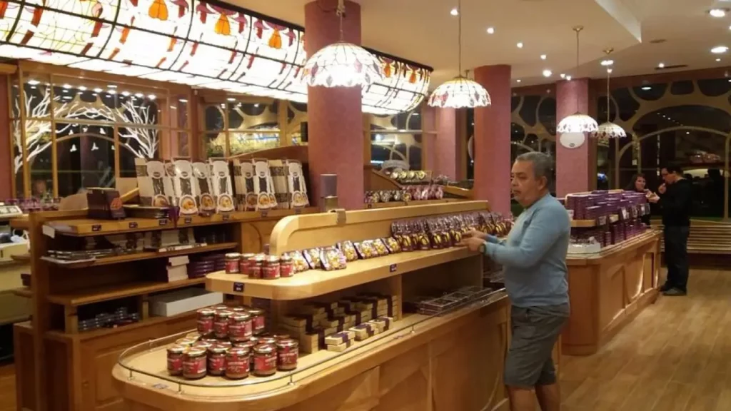 Interior de uma loja de doces ou chocolates finos com prateleiras de madeira, iluminação aconchegante e um homem escolhendo produtos embalados.